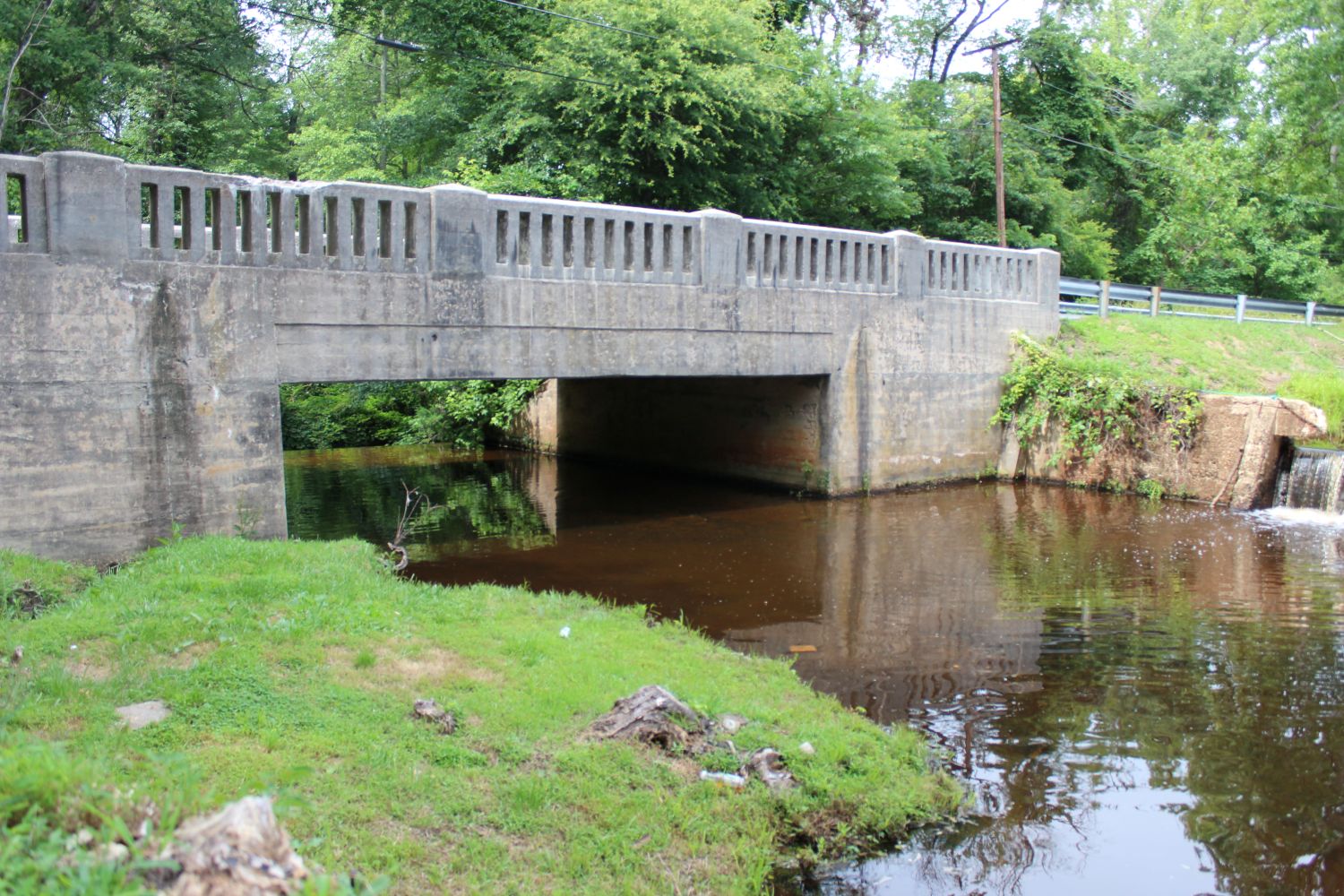 The Bridge and Spillway Union: The concrete bridge overpass channels the dark, tannin-stained waters of the Deep Run system toward the main overflow mechanism. To the far right, a dedicated notch in the concrete infrastructure funnels a steady, controlled cascade, protecting the structural integrity of the bypass channel.  