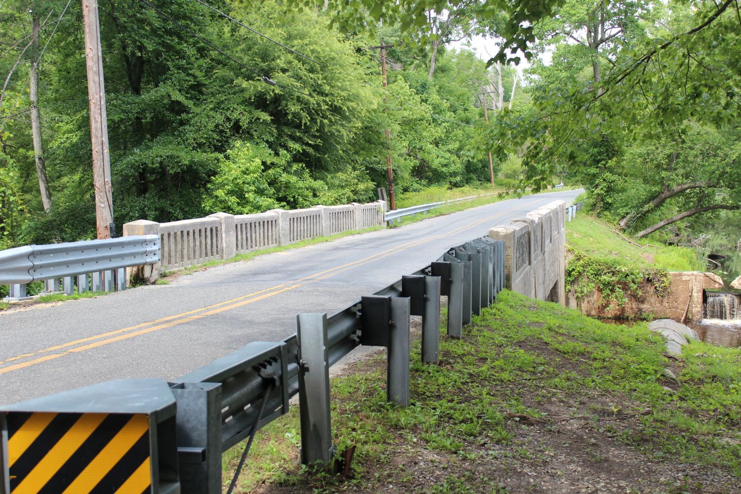 Waterworks Road Crossing: A roadside perspective along Waterworks Road as it spans across the Elkinton Pond Dam infrastructure. Flanked by modern metal guardrails and historic concrete balustrades, this rural pass sits directly atop the hydrological boundary separating the main body of the pond from its downstream runoff channel. 