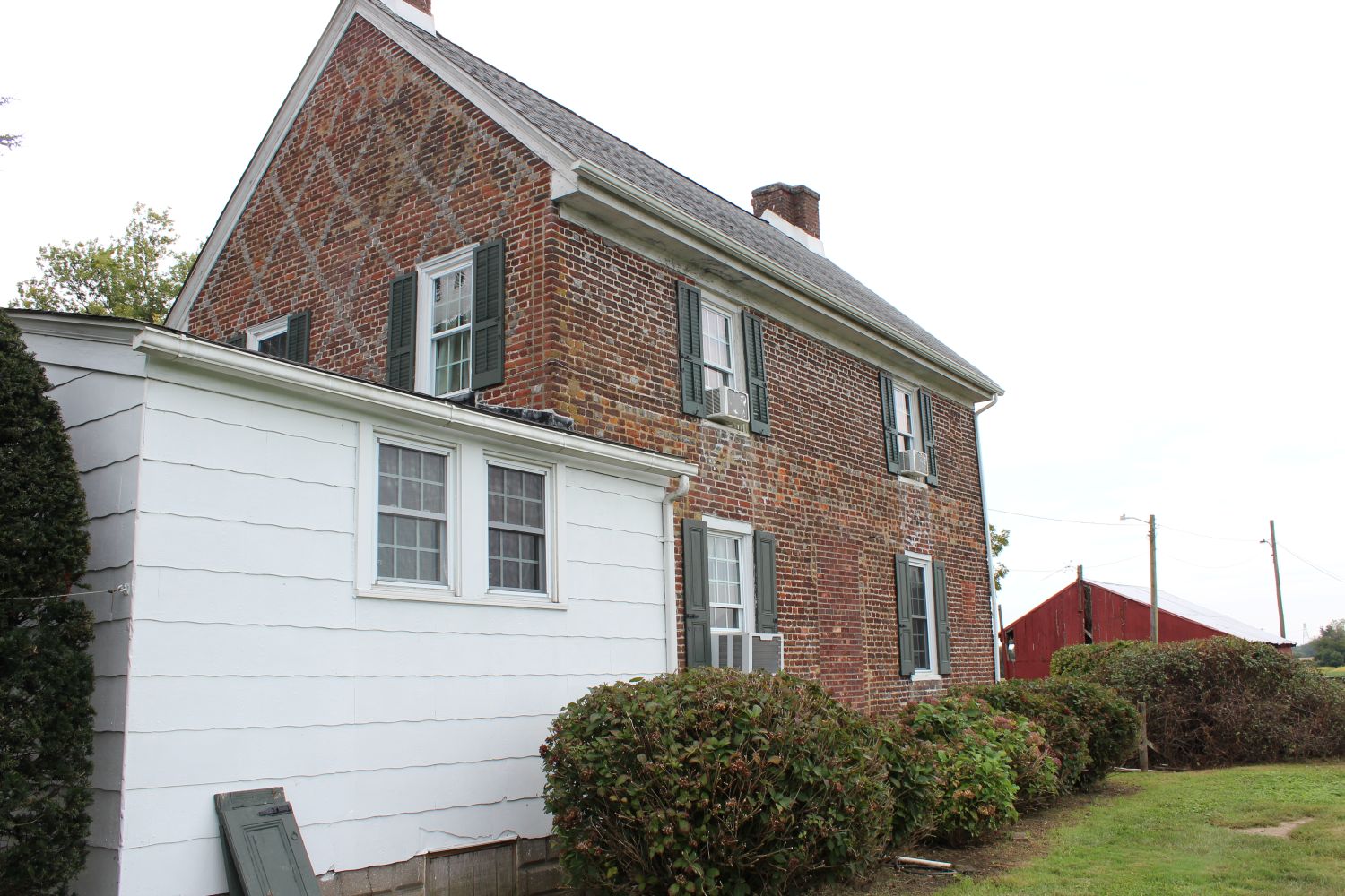 Two-story brick house with green shutters and a white one-story addition, seen from the side, diamond brick pattern faintly visible on the gable end.