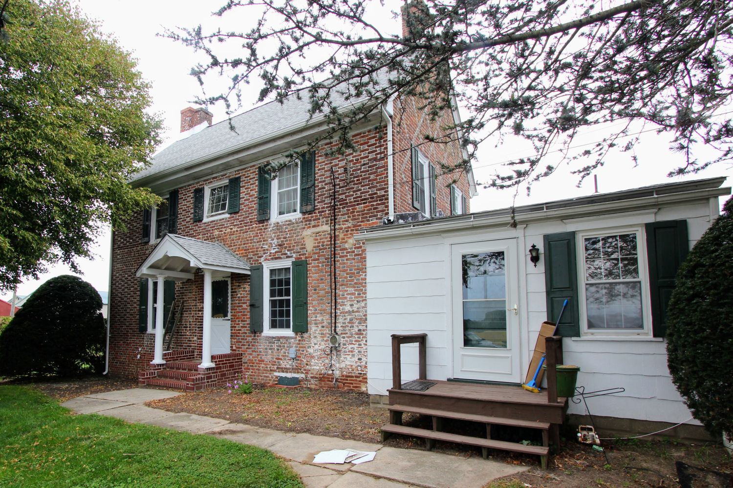 Front view of a brick house's white entry portico with columns, green shutters, and a ladder leaning against the wall, seen through tree branches.
