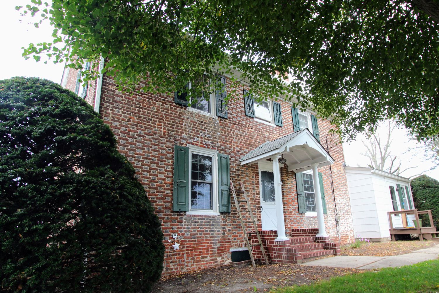 Front view of a brick house's white entry portico and green shutters, partly shaded by an overhanging tree.