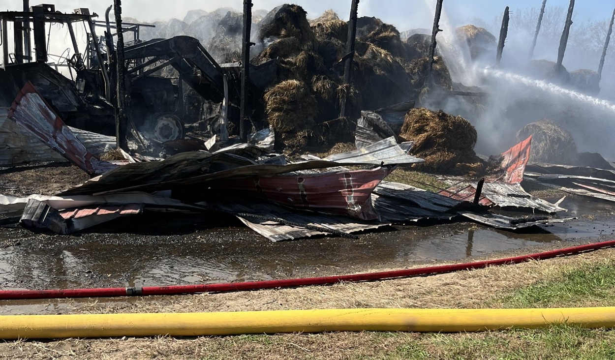 Burned tractors and smoldering hay bales sit among the charred barn posts as streams of water reach the pile.